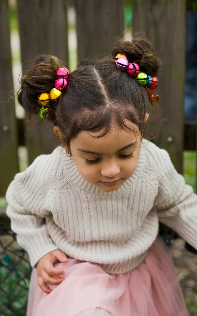 Capelli da bambino raccolti in doppi chignon con accessori colorati jingle bell per un look natalizio' title=
