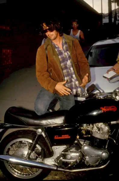 Pozat în jurul anului 1991, Keanu Reeves poartă o jachetă din shearling cu nasturi în carouri în timp ce se urcă pe motocicleta sa.