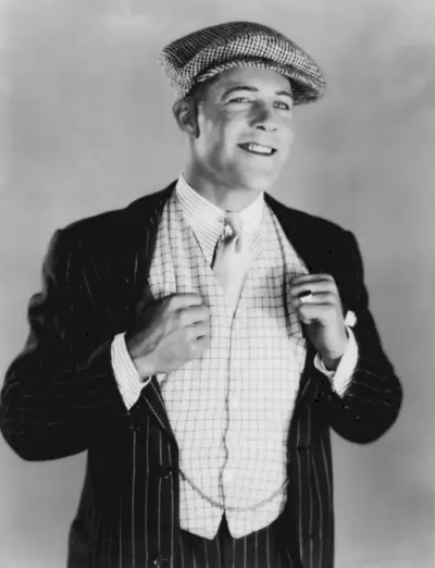 Aktor Kenneth Harlan ma na sobie stylową kamizelkę z garniturem w prążki, koszulę w paski i szpilkę do kołnierzyka oraz płaską czapkę.