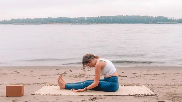 Woman sitting on a yoga mat at the beach leaning her chest toward her thighs in a Yin Yoga pose