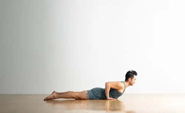 Man with dark hair practices Cobra Pose on a wood floor. The background is white. He is wearing light blue clothes.