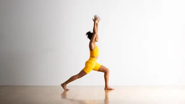 A brown-skinned woman wearing a bright yellow top and shorts, practices High Lunge with her arms extended up