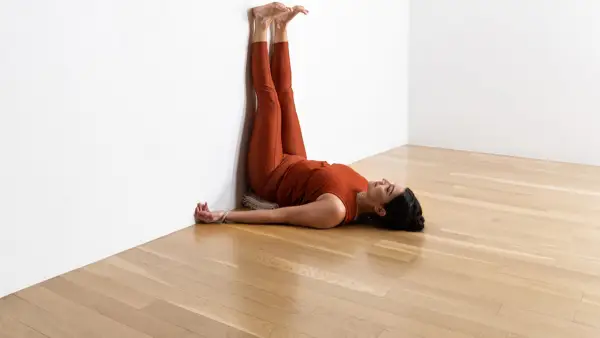A woman with dark hair, wearing a copper colored tights and top practices Legs Up the Wall Pose. She is lying on a wood floor with her legs extended up a white wall.