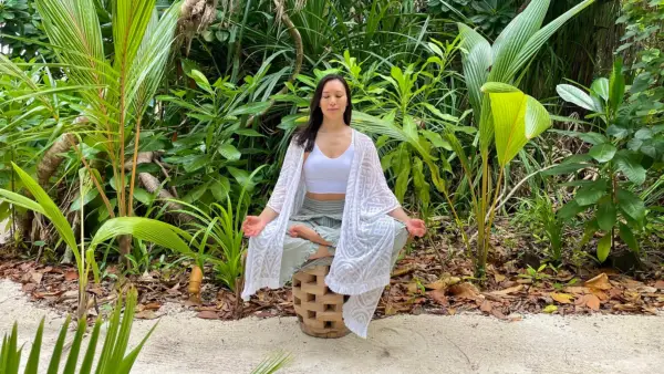 Ingrid Yang sits on a small bench in the sand, surrounded by tropical foliage. She is wearing a translucent white wrap and loose, light green pants.