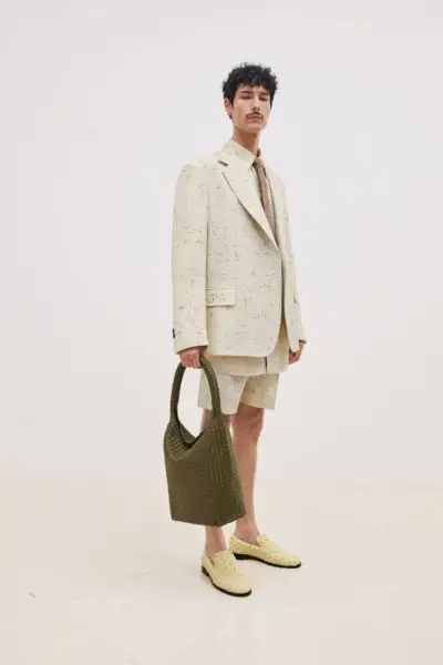 Noah Ramos fa una figura affascinante con un abito corto color crema screziato con cravatta coordinata, accessoriato con mocassini intrecciati e una borsa tote verde oliva, che incarna Bottega Veneta
