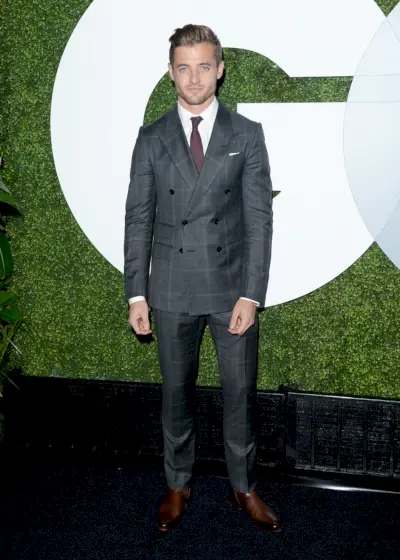 Robbie Rogers indossa un abito New Signoria doppiopetto scozzese grigio con revers a lancia con camicia bianca, cravatta rossa e stivali di pelle marroni.