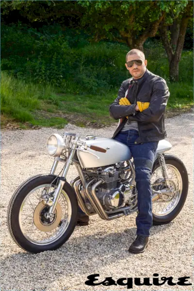 Assis sur un vélo, Tom Hardy porte une veste en cuir et un jean Belstaff. Il porte également un t-shirt Alternative Apparel, des bottes Triumph et des lunettes de soleil Carrera.