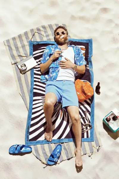Seth Rogen slapper af på et Hermès strandhåndklæde og bærer en Dior Men-skjorte, Calvin Klein tank, POLO Ralph Lauren-shorts, Birkenstock-sandaler og Garrett Leight-solbriller.