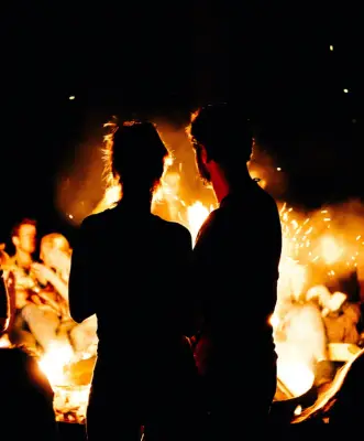 Silhouette of a couple standing in front of a glowing yellow fire with other people sitting around the fire.