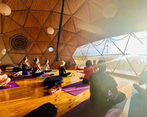A group of people sit cross-legged on yoga mats on a wood floor. They are sitting in a geodesic dome in Patagonia. The sun set streams in