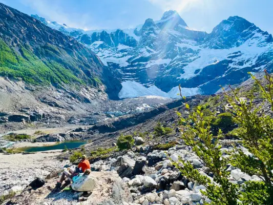 People rest along a rocky hiking trail in the Patagonia. In the foreground are rocks and green brush. Below them is a pool of deep blue green water, and in the background green hills rise up into the snow dusted mountains. 