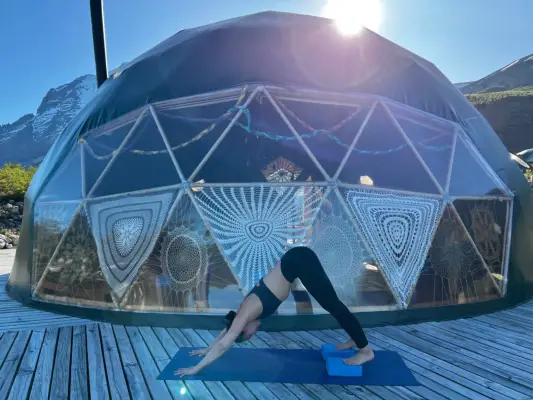 A woman practices Downward Facing dog on a blue mat, set on a deck in front of a geodesic dome. Some of the triangular windows have crocheted curtains covering them. Located in Patagonia.