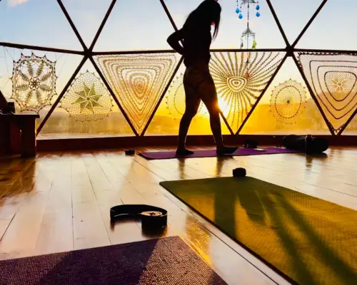 A woman is silhouetted against the triangular windows of a geodesic dome. In the foreground there are yoga mats rolled out.