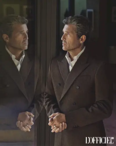 Patrick Dempsey, som spejler sig i glasset, er klædt i en tekstureret brun frakke, der taler til tidløs elegance.