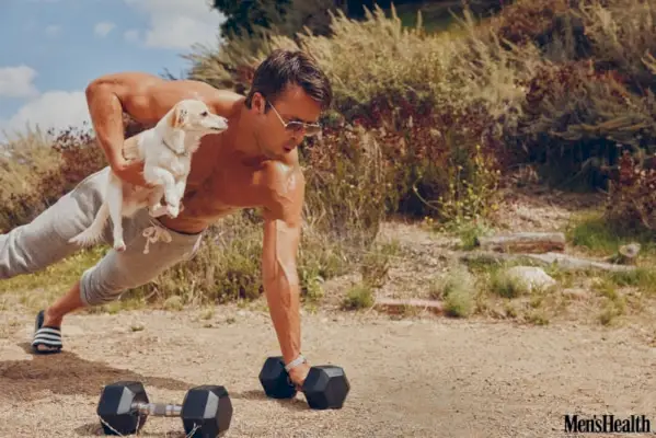 Glen Powell izvaja sklece z eno roko na prostem, medtem ko uravnoteži svojega psa Brisketa. Nosi sončna očala Persol s trenirkami Todd Snyder x Champion in uro OMEGA.