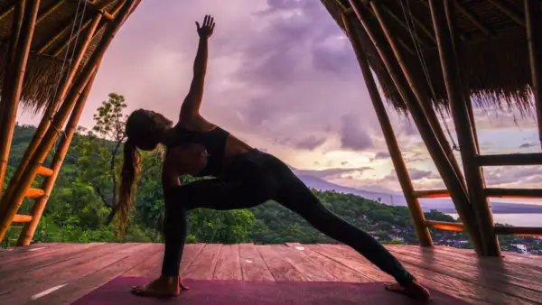 woman doing extended triangle pose on yoga mat outside under thatched room with trees in the distance